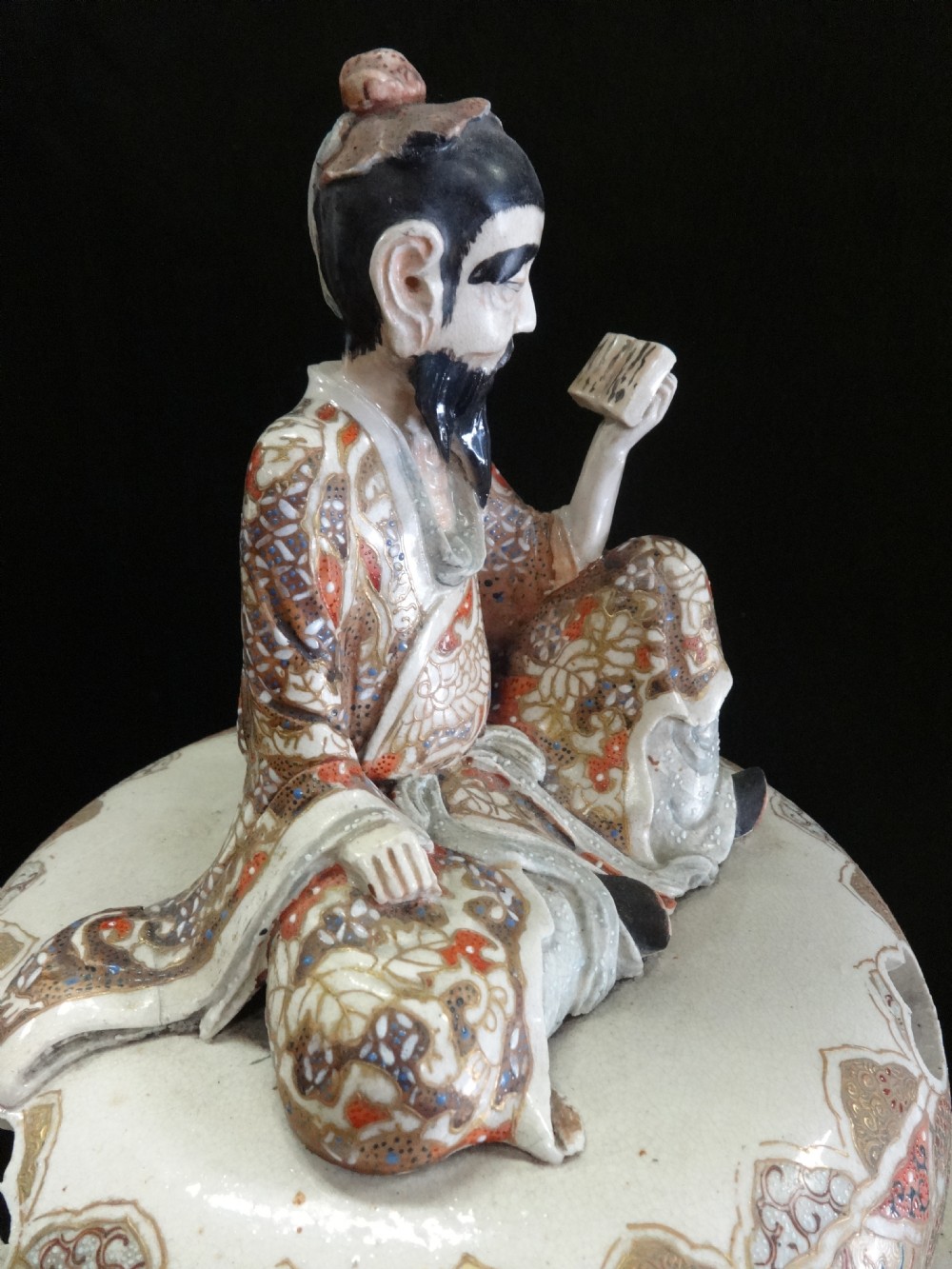 An antique Japanese Satsuma ceramic koro incense burner featuring tripodic legs, side handles, hand-painted figures, gold embellishments, and a seated figure finial on the lid. View VII