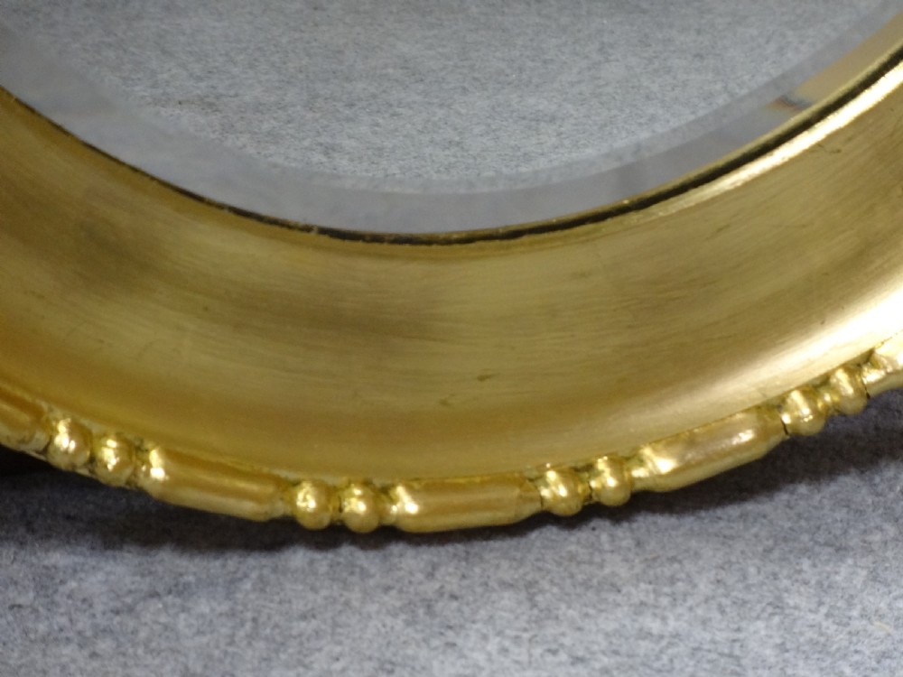 An oval Edwardian wall mirror with a gilded frame featuring beaded trim details around a bevelled glass mirror plate. View V