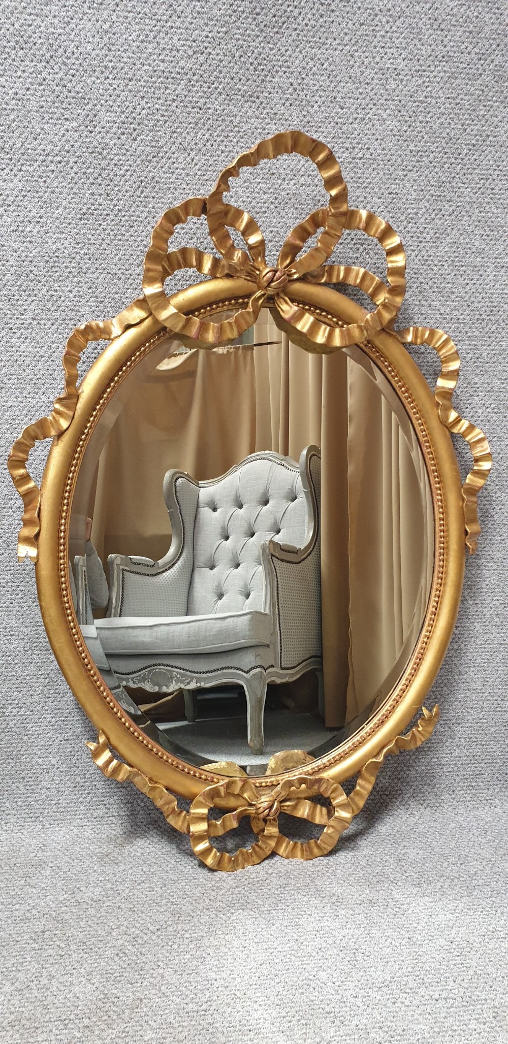 An oval giltwood wall mirror featuring carved ribbon bow detailing at the top and base with a beaded inner rim.