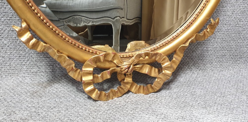 An oval giltwood wall mirror featuring carved ribbon bow detailing at the top and base with a beaded inner rim. View III