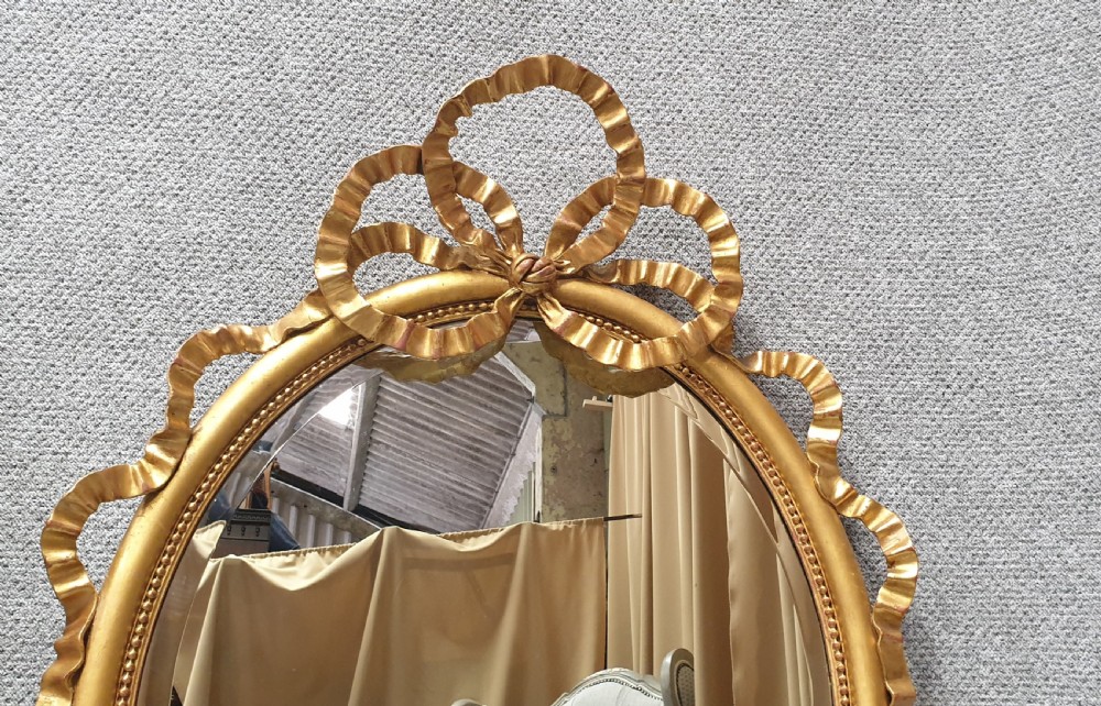 An oval giltwood wall mirror featuring carved ribbon bow detailing at the top and base with a beaded inner rim. View IV