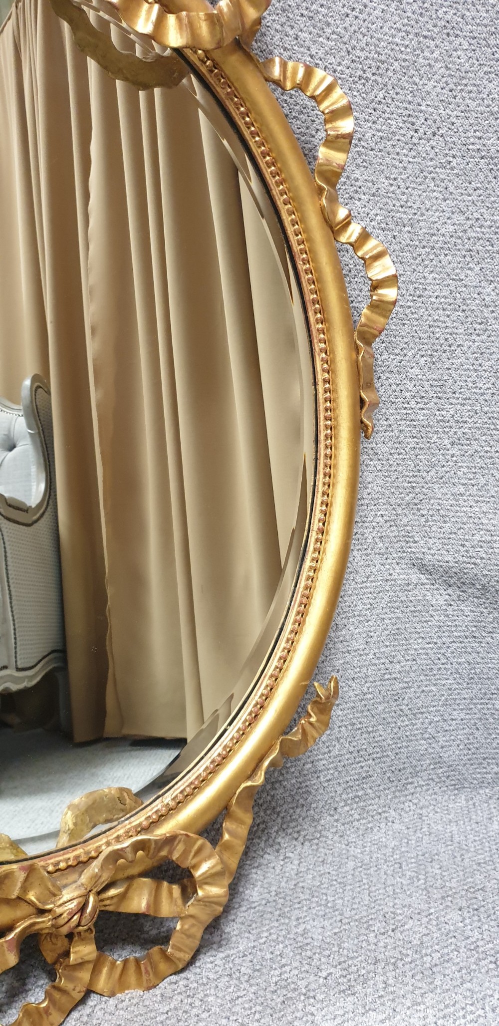 An oval giltwood wall mirror featuring carved ribbon bow detailing at the top and base with a beaded inner rim. View V