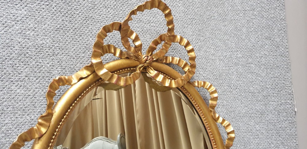 An oval giltwood wall mirror featuring carved ribbon bow detailing at the top and base with a beaded inner rim. View VI
