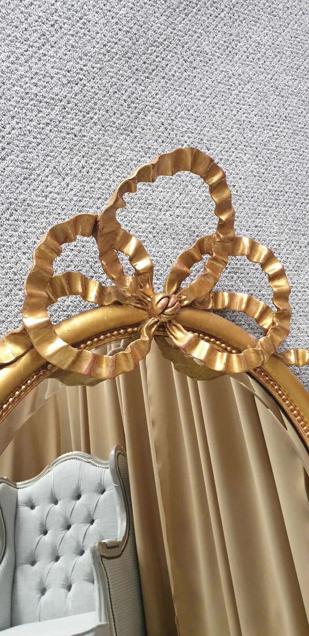 An oval giltwood wall mirror featuring carved ribbon bow detailing at the top and base with a beaded inner rim. View VII