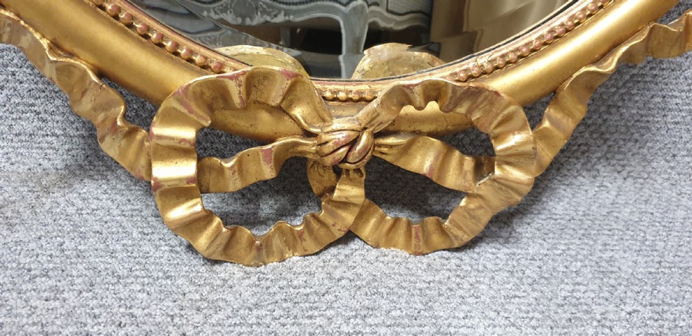 An oval giltwood wall mirror featuring carved ribbon bow detailing at the top and base with a beaded inner rim. View VIII