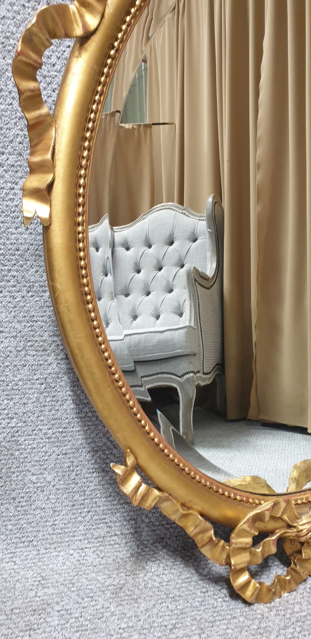An oval giltwood wall mirror featuring carved ribbon bow detailing at the top and base with a beaded inner rim. View IX