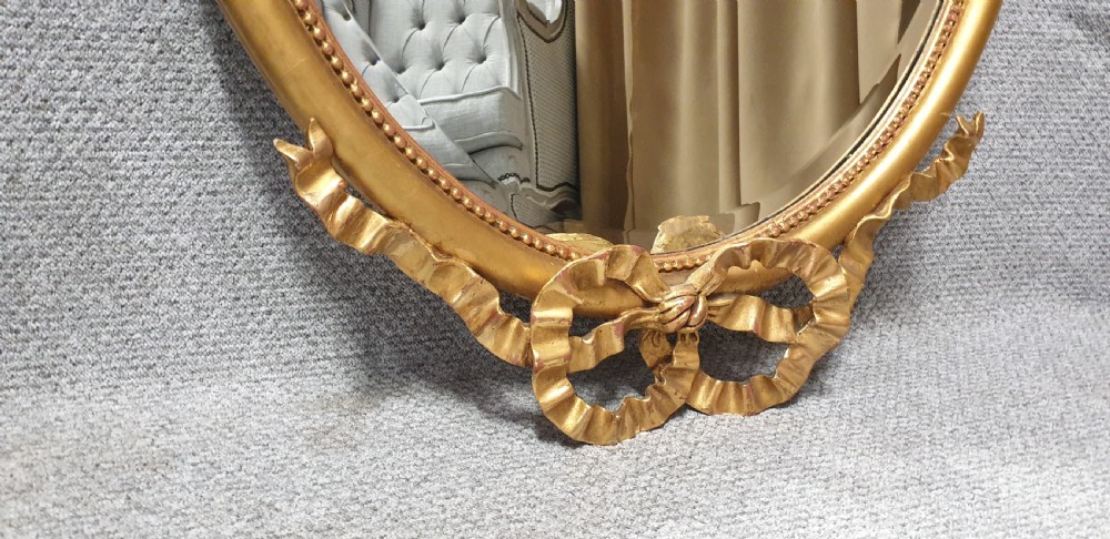 An oval giltwood wall mirror featuring carved ribbon bow detailing at the top and base with a beaded inner rim. View X
