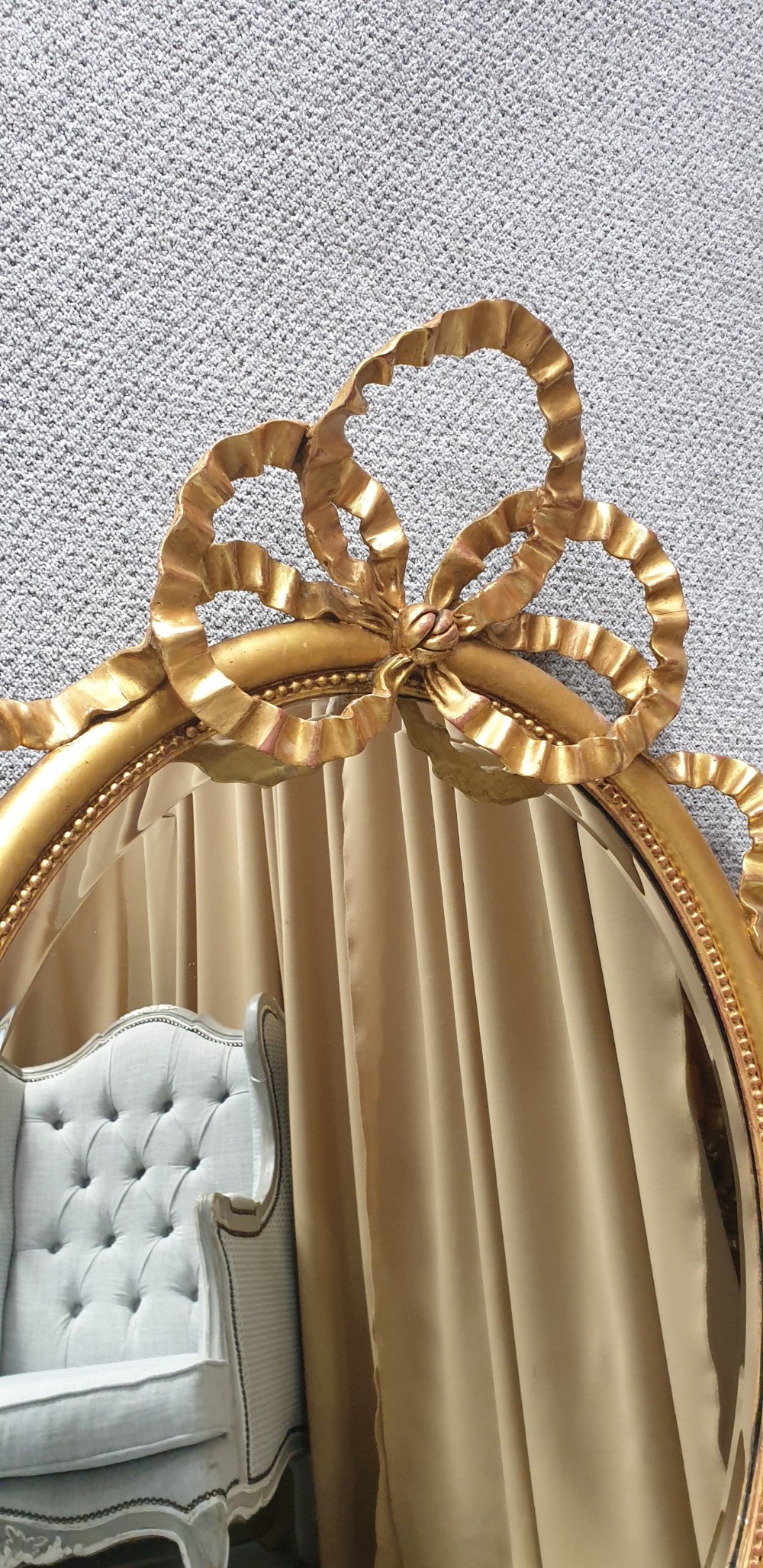 An oval giltwood wall mirror featuring carved ribbon bow detailing at the top and base with a beaded inner rim. View XI