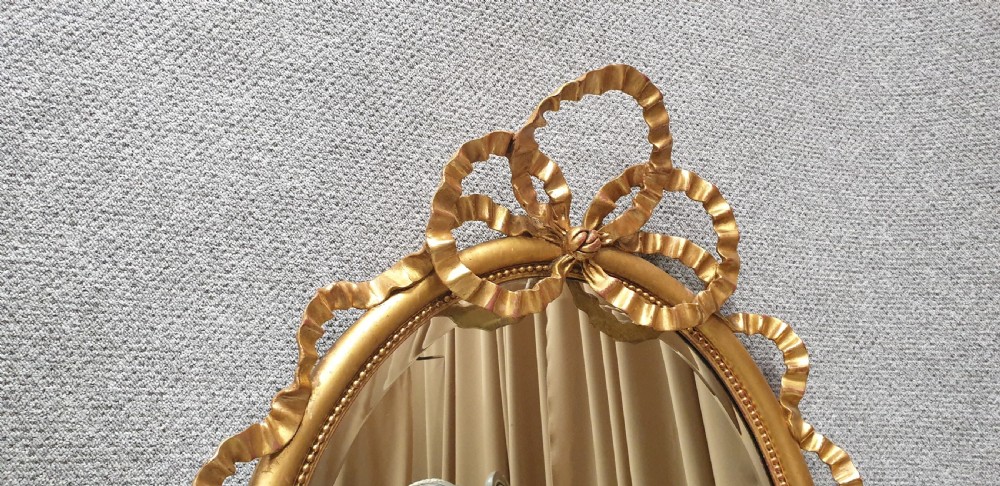 An oval giltwood wall mirror featuring carved ribbon bow detailing at the top and base with a beaded inner rim. View XIII