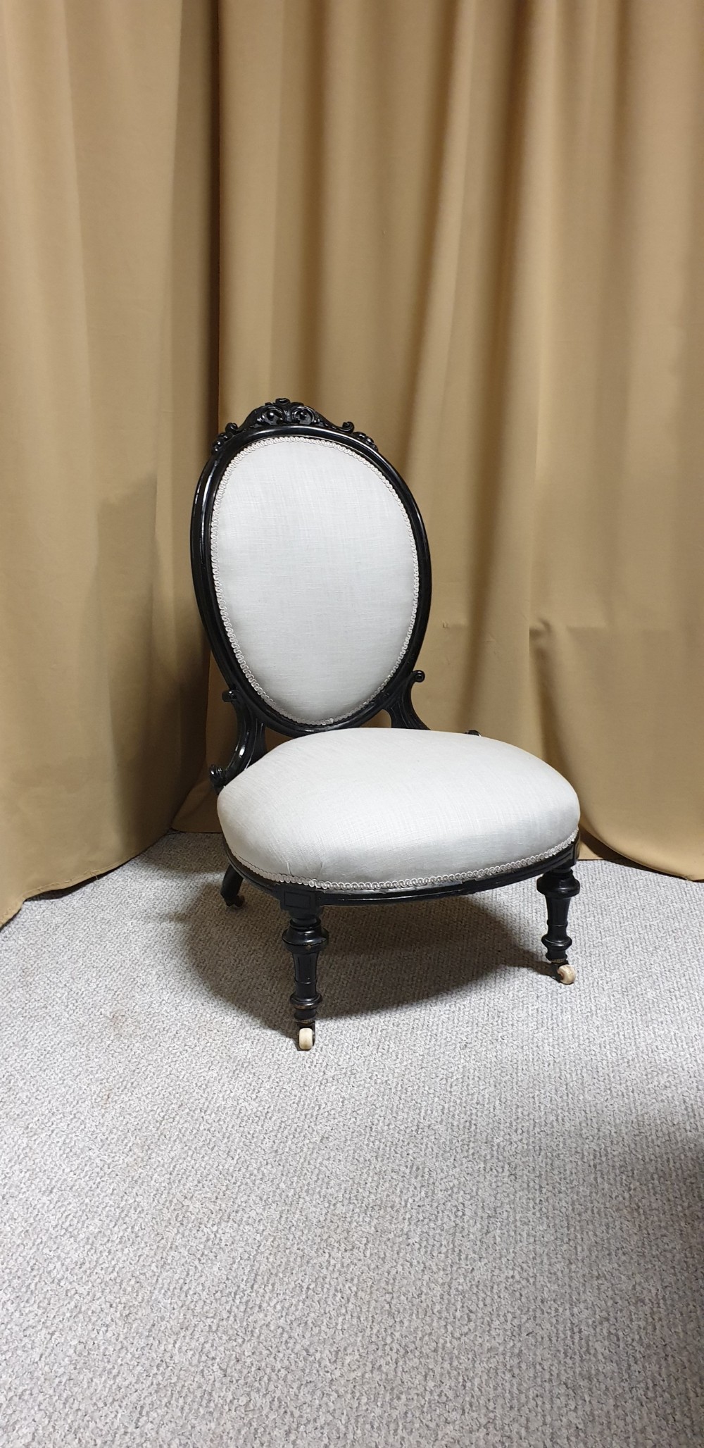 A Victorian low-slung nursing chair featuring an ebonised black wood frame with an oval backrest, carved top crest, cream upholstered seat, and turned front legs with castors.