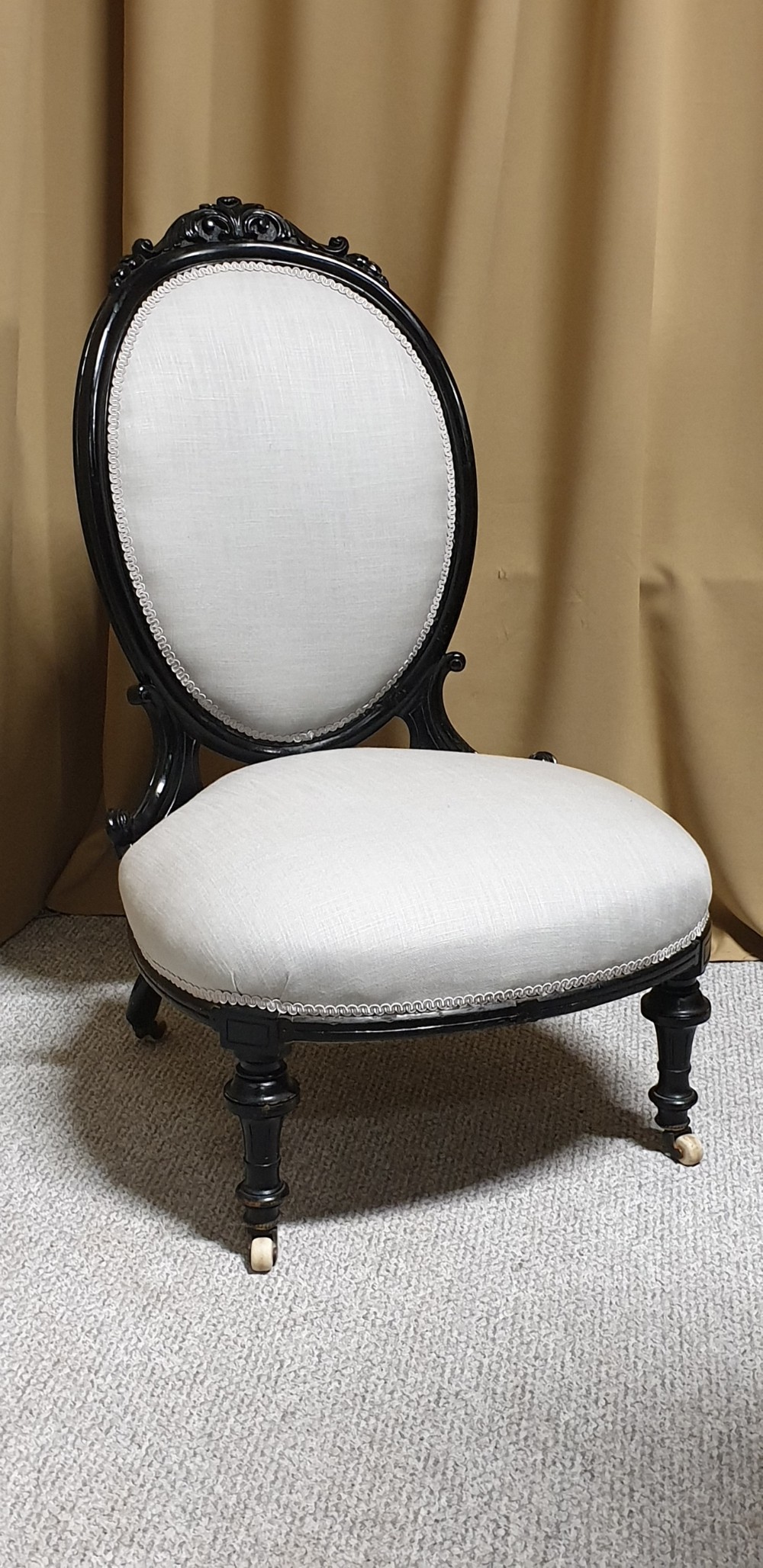 A Victorian low-slung nursing chair featuring an ebonised black wood frame with an oval backrest, carved top crest, cream upholstered seat, and turned front legs with castors. View II