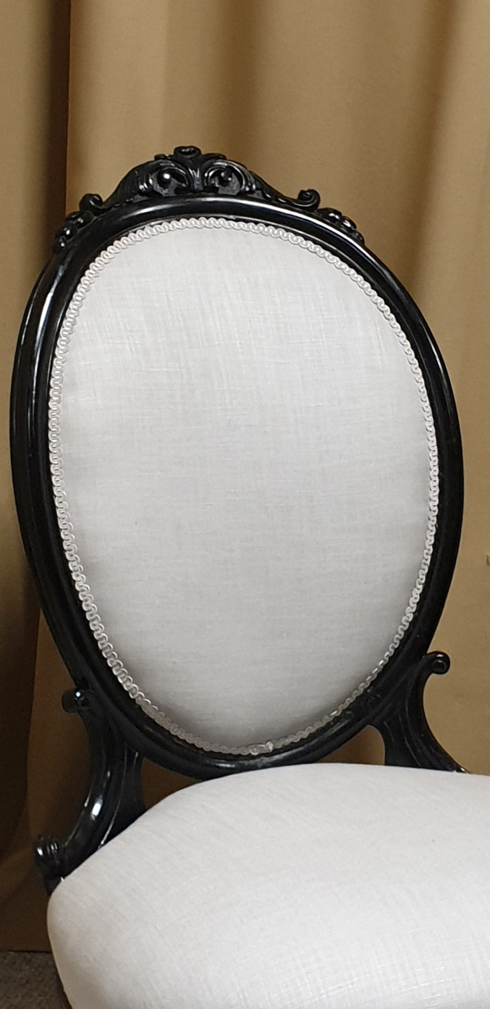 A Victorian low-slung nursing chair featuring an ebonised black wood frame with an oval backrest, carved top crest, cream upholstered seat, and turned front legs with castors. View III