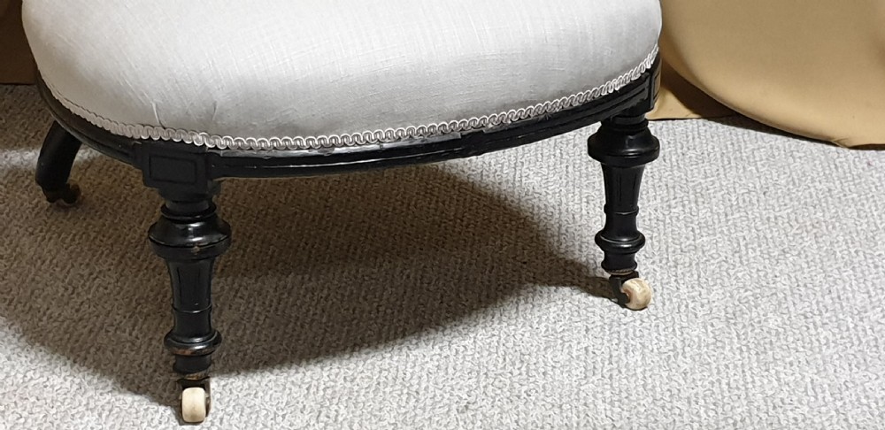 A Victorian low-slung nursing chair featuring an ebonised black wood frame with an oval backrest, carved top crest, cream upholstered seat, and turned front legs with castors. View IV