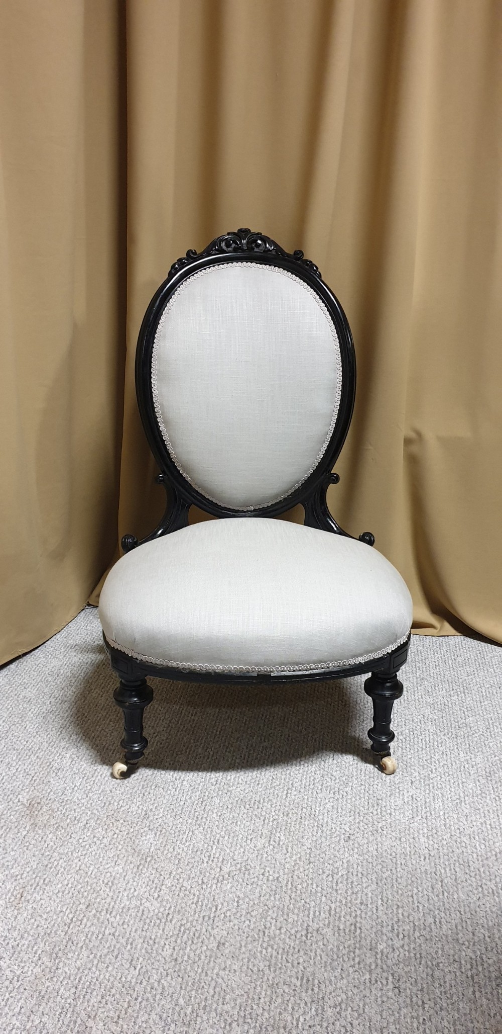 A Victorian low-slung nursing chair featuring an ebonised black wood frame with an oval backrest, carved top crest, cream upholstered seat, and turned front legs with castors. View V