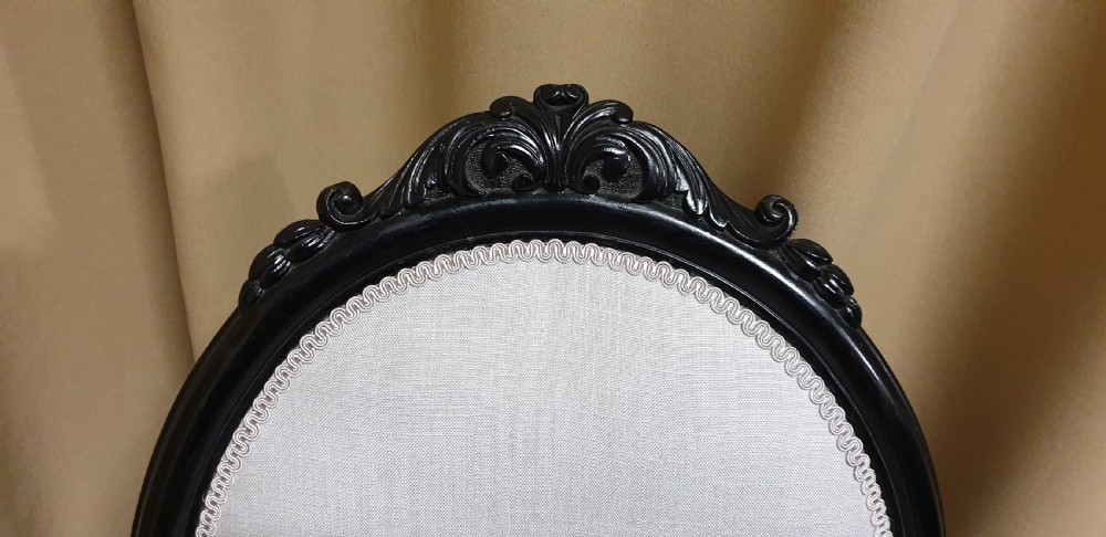 A Victorian low-slung nursing chair featuring an ebonised black wood frame with an oval backrest, carved top crest, cream upholstered seat, and turned front legs with castors. View VI