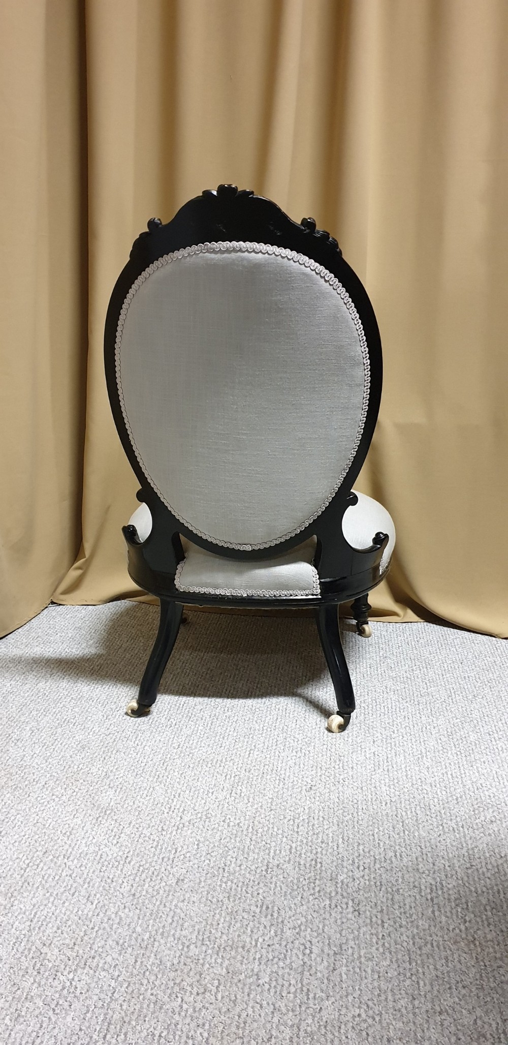 A Victorian low-slung nursing chair featuring an ebonised black wood frame with an oval backrest, carved top crest, cream upholstered seat, and turned front legs with castors. View VII