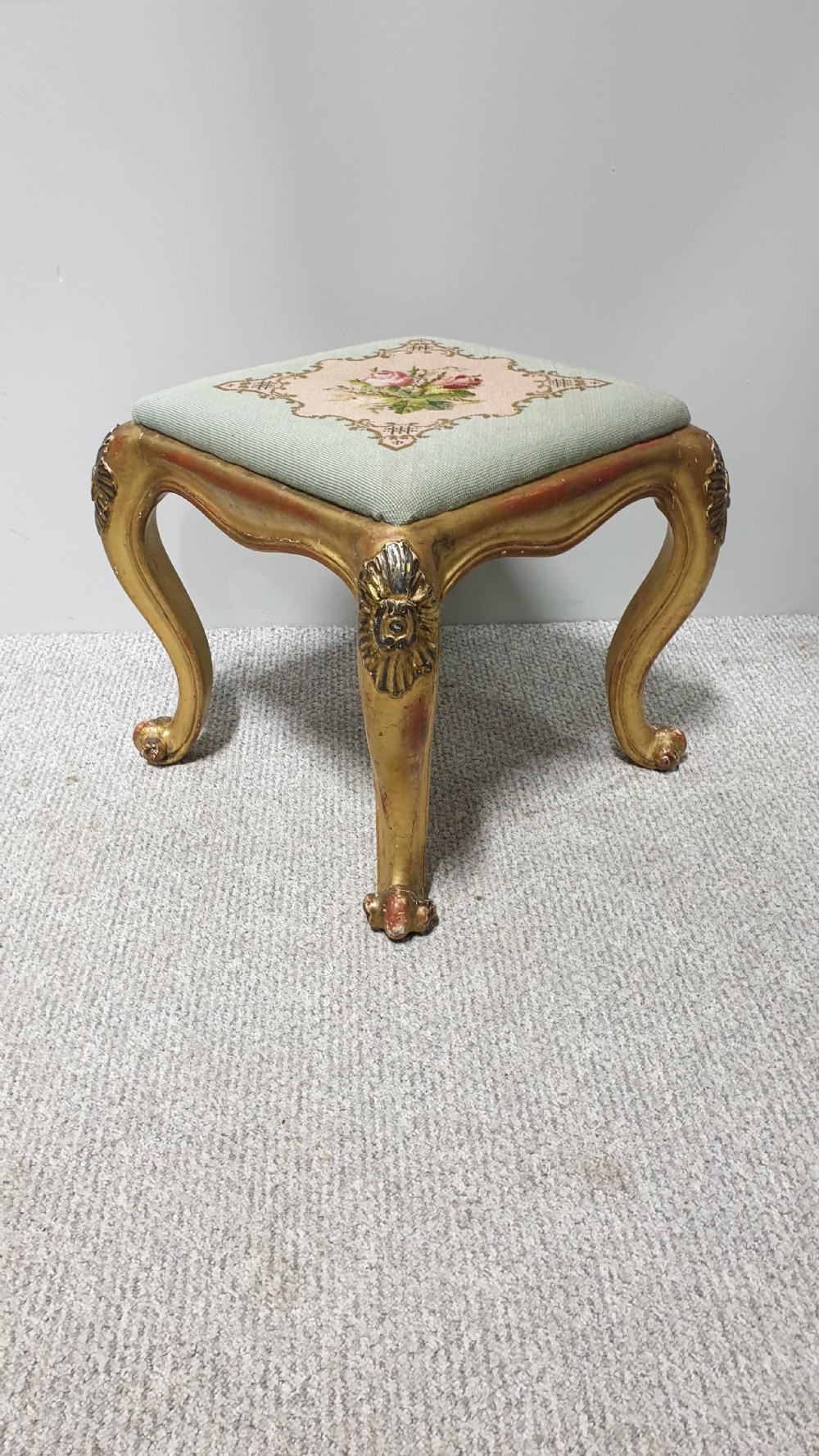 A square Victorian giltwood footstool featuring carved cabriole legs and a needlepoint floral seat on a pale green background.