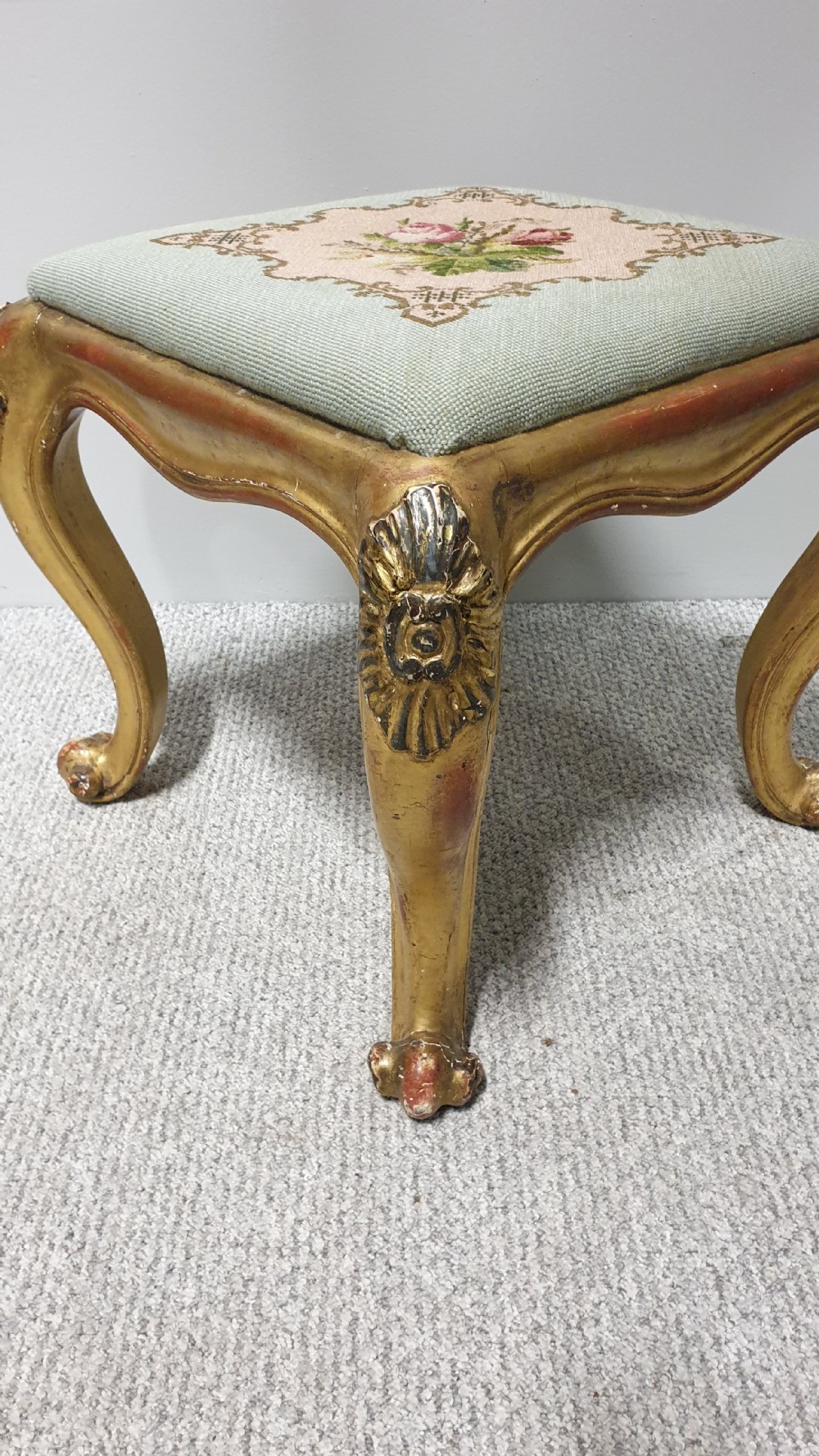 A square Victorian giltwood footstool featuring carved cabriole legs and a needlepoint floral seat on a pale green background. View II