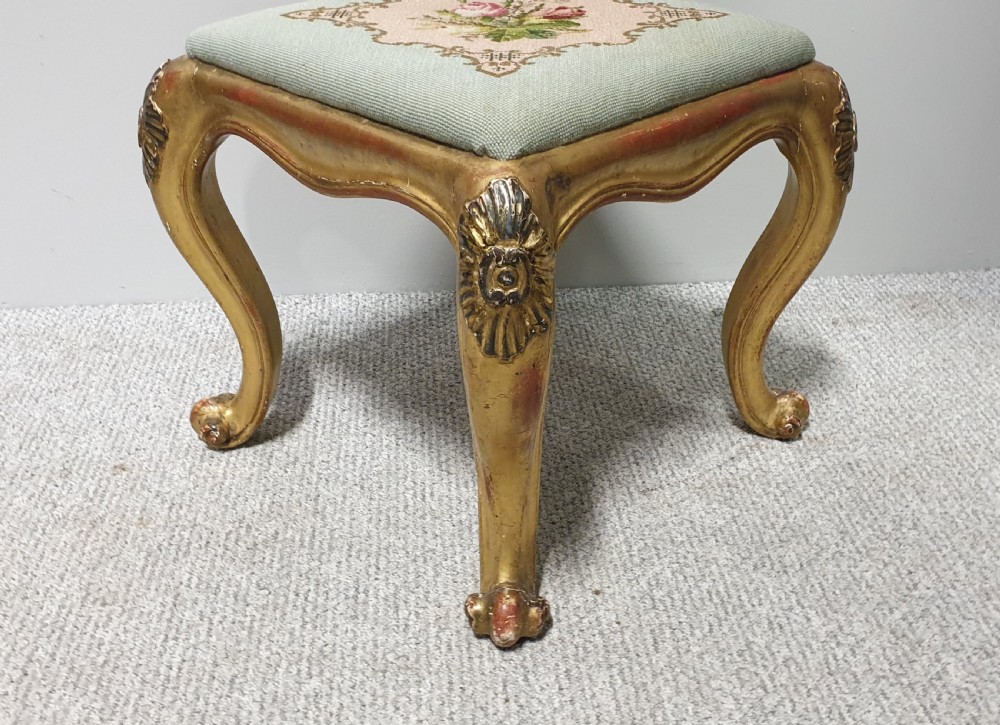 A square Victorian giltwood footstool featuring carved cabriole legs and a needlepoint floral seat on a pale green background. View III