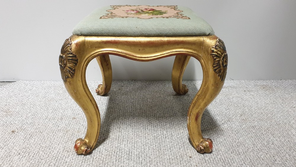 A square Victorian giltwood footstool featuring carved cabriole legs and a needlepoint floral seat on a pale green background. View IV