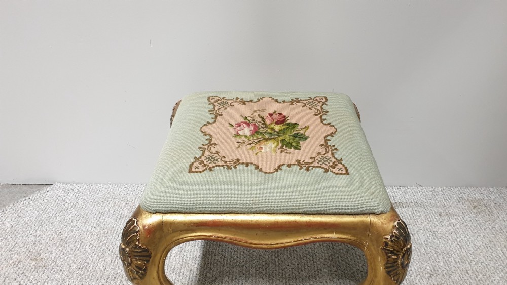 A square Victorian giltwood footstool featuring carved cabriole legs and a needlepoint floral seat on a pale green background. View V