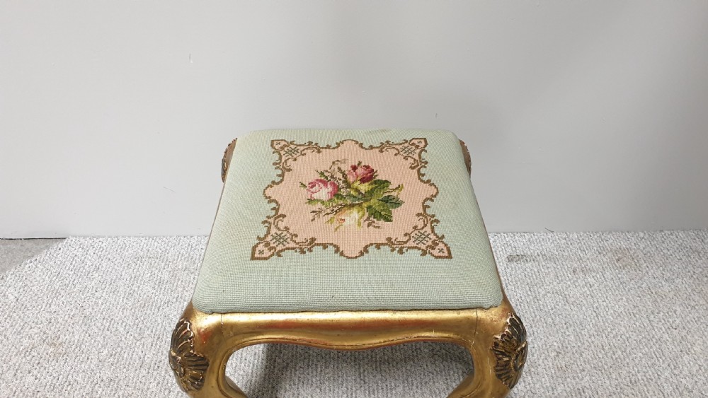 A square Victorian giltwood footstool featuring carved cabriole legs and a needlepoint floral seat on a pale green background. View VI