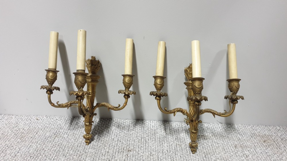 A pair of French Neoclassical gilt brass three-light wall sconces featuring fluted torch backplates, scrolling foliate arms, and cream candle sleeves.