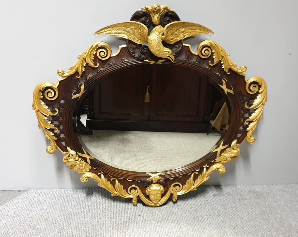 An ornate oval mahogany wall mirror featuring a carved eagle crest, parcel gilt acanthus leaf borders, side lattice panels, and a central classical mask motif at the base.
