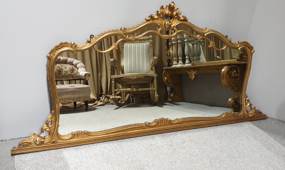 A wide French Rococo style gilt overmantel mirror with an ornate carved central crest and sweeping serpentine gold frame.