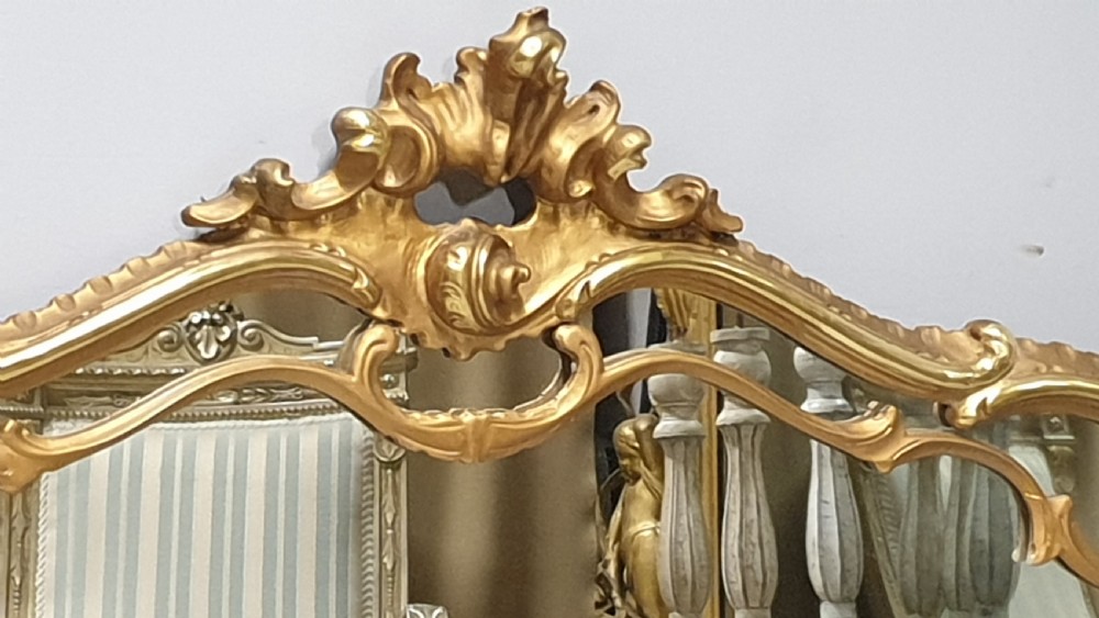 A wide French Rococo style gilt overmantel mirror with an ornate carved central crest and sweeping serpentine gold frame. View III