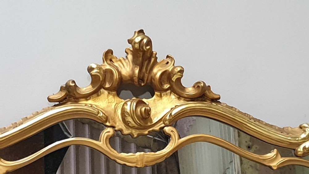 A wide French Rococo style gilt overmantel mirror with an ornate carved central crest and sweeping serpentine gold frame. View VI
