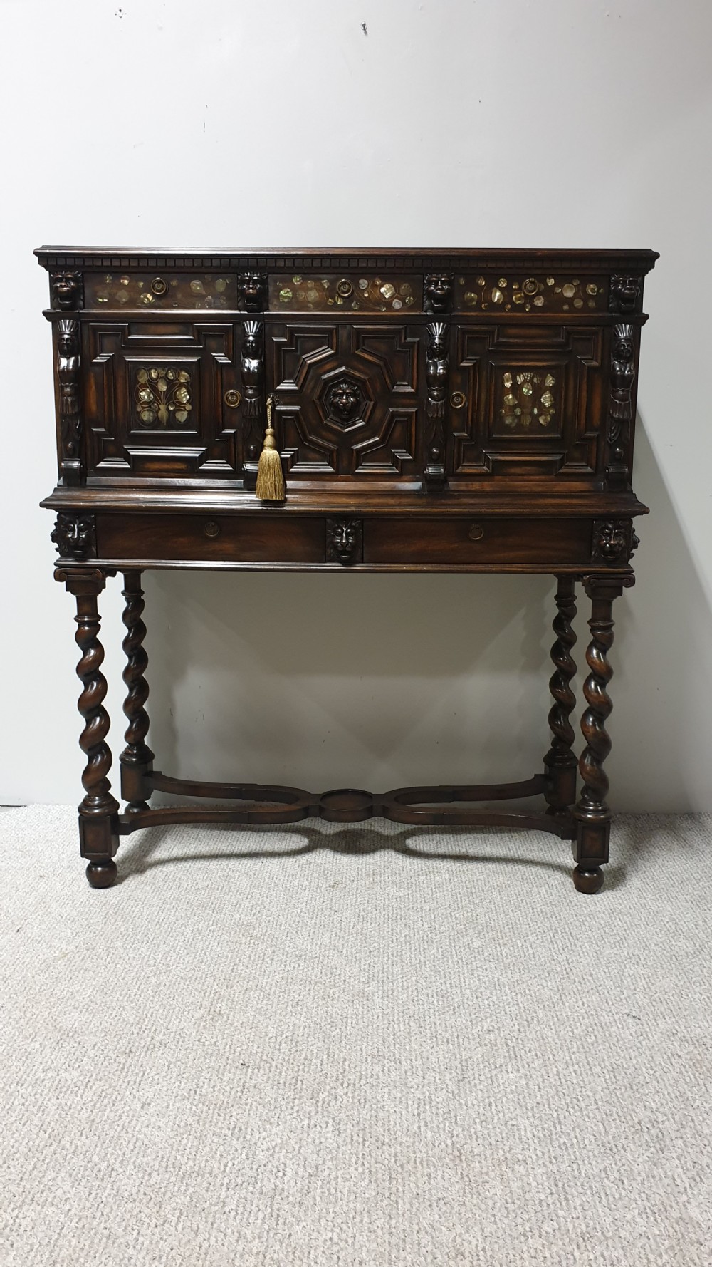 A dark carved walnut cabinet on a stand featuring mother of pearl inlay, carved lion mask details, geometric door panels, and barley twist legs with a stretcher.