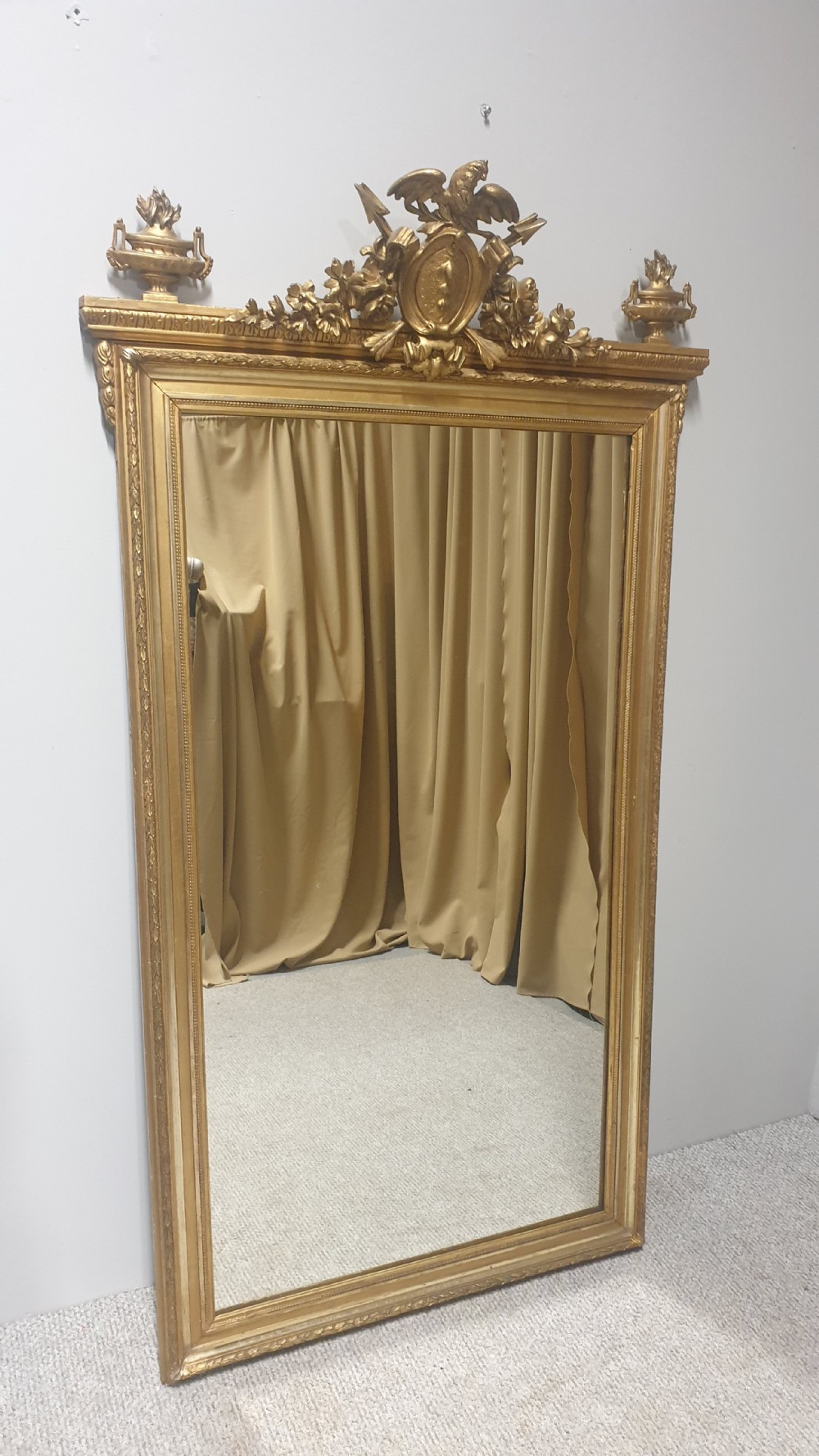 A tall rectangular French giltwood mirror featuring an ornate eagle crest flanked by urn finials and moulded frame detailing.