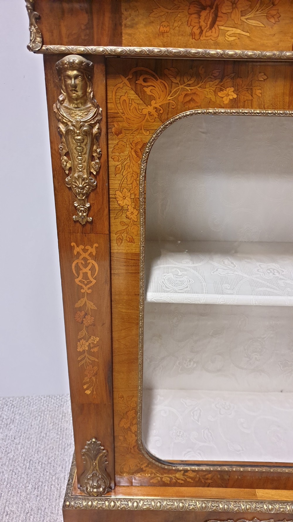A Victorian walnut pier cabinet featuring floral marquetry inlays, gilt metal ormolu mounts, and a single glazed door opening to light-coloured shelves. View VI