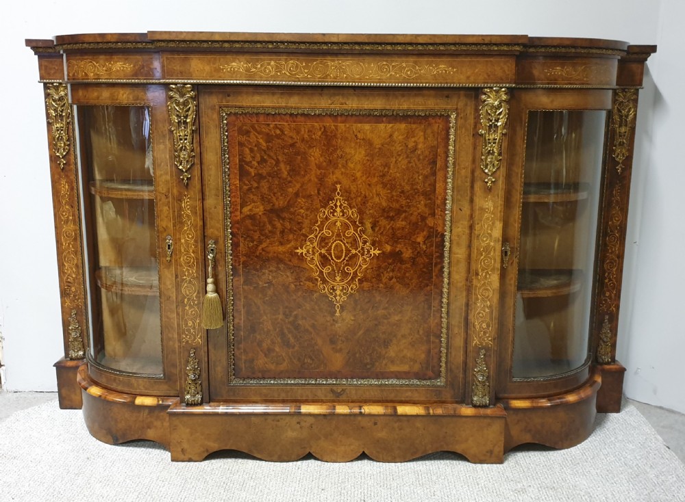 A Victorian burr walnut credenza featuring a central inlaid cupboard door flanked by two curved glazed display side cabinets and decorative gilt ormolu mounts.