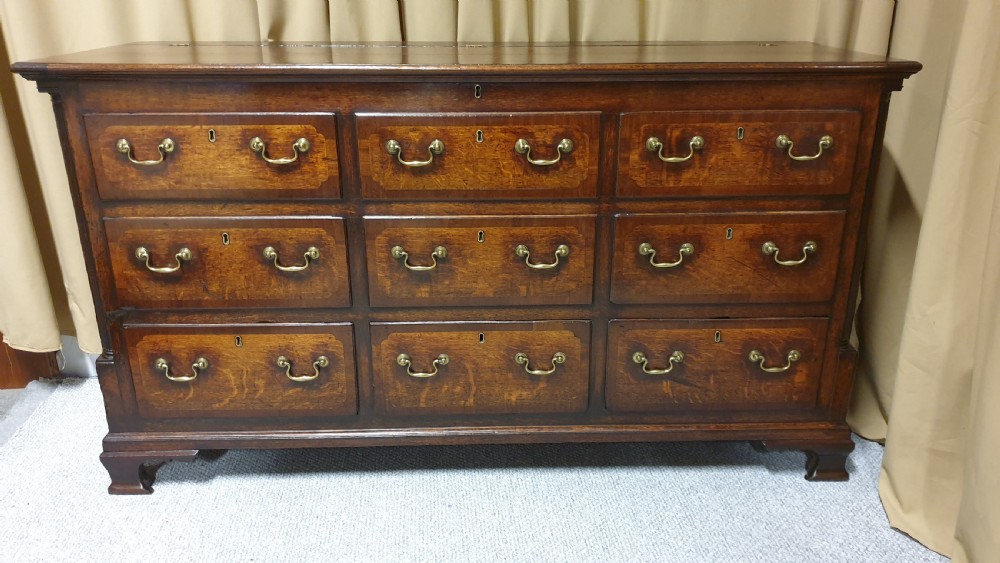 A dark oak Georgian Lancashire coffer featuring nine fielded panel drawer fronts with brass swan-neck handles and a hinged top lid resting on ogee bracket feet. View II
