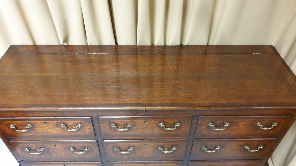 A dark oak Georgian Lancashire coffer featuring nine fielded panel drawer fronts with brass swan-neck handles and a hinged top lid resting on ogee bracket feet. View VII
