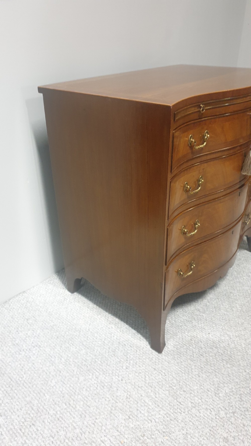 A small mahogany chest of drawers featuring a curved serpentine front, four graduated drawers with brass handles, and bracket feet. View V