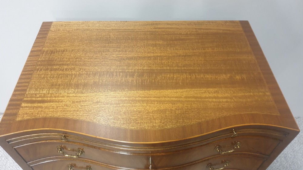 A small mahogany chest of drawers featuring a curved serpentine front, four graduated drawers with brass handles, and bracket feet. View VI