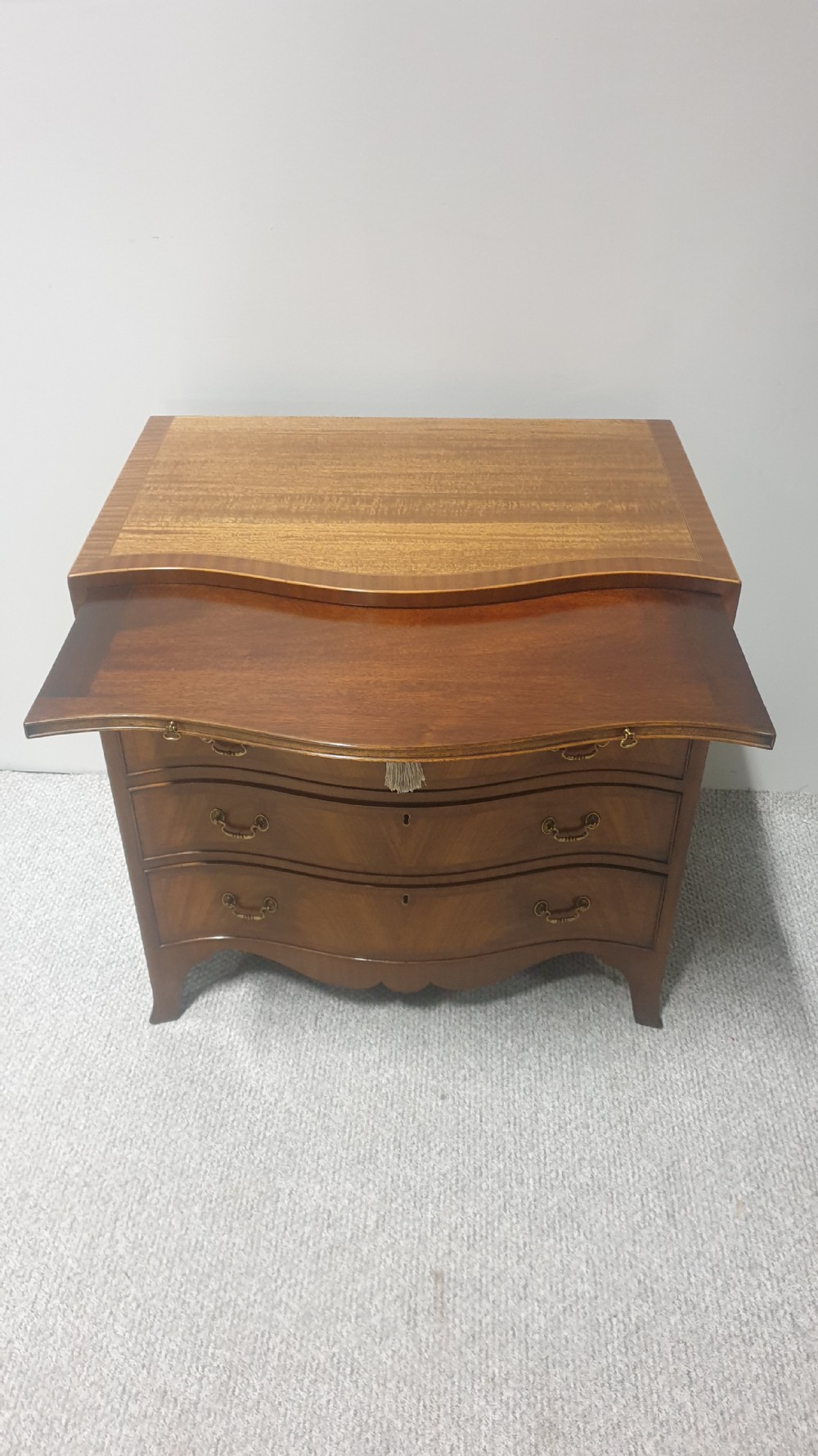 A small mahogany chest of drawers featuring a curved serpentine front, four graduated drawers with brass handles, and bracket feet. View VII