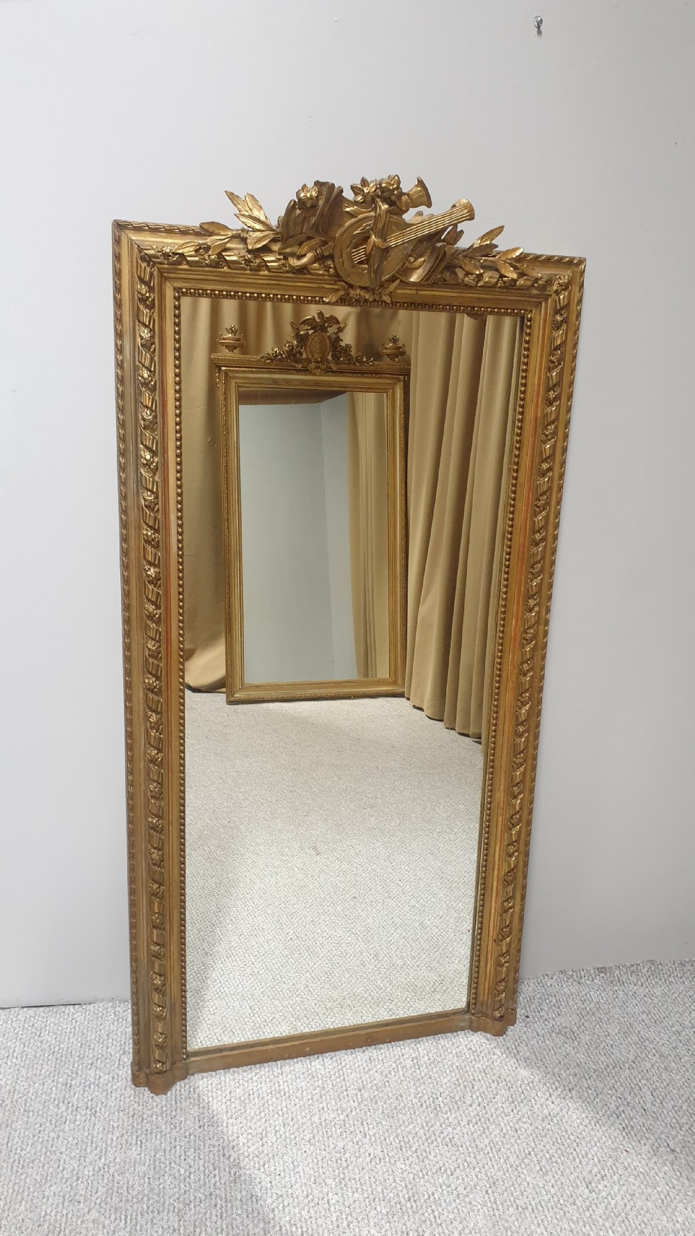A tall rectangular French giltwood pier mirror topped with an ornate carved crest featuring a musical lyre and foliage.