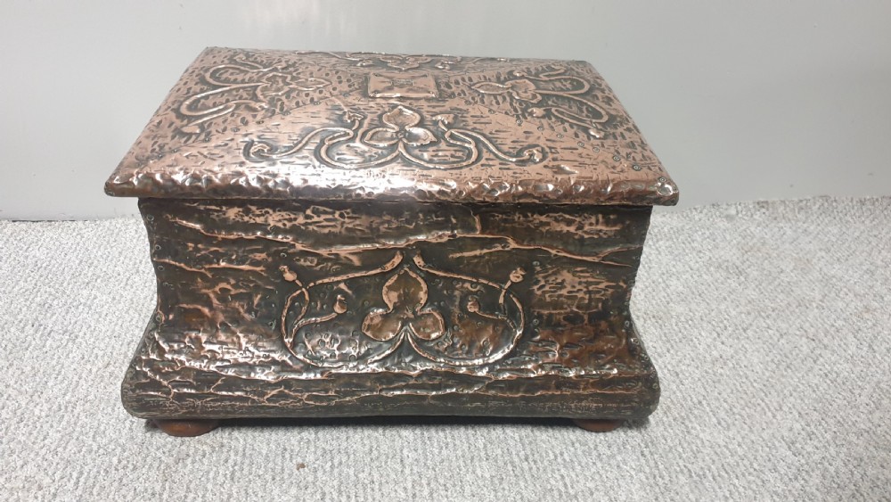 An antique rectangular copper fireside box featuring a hinged lid, hammered repousse foliate motifs, flared sides, and small bun feet. View III