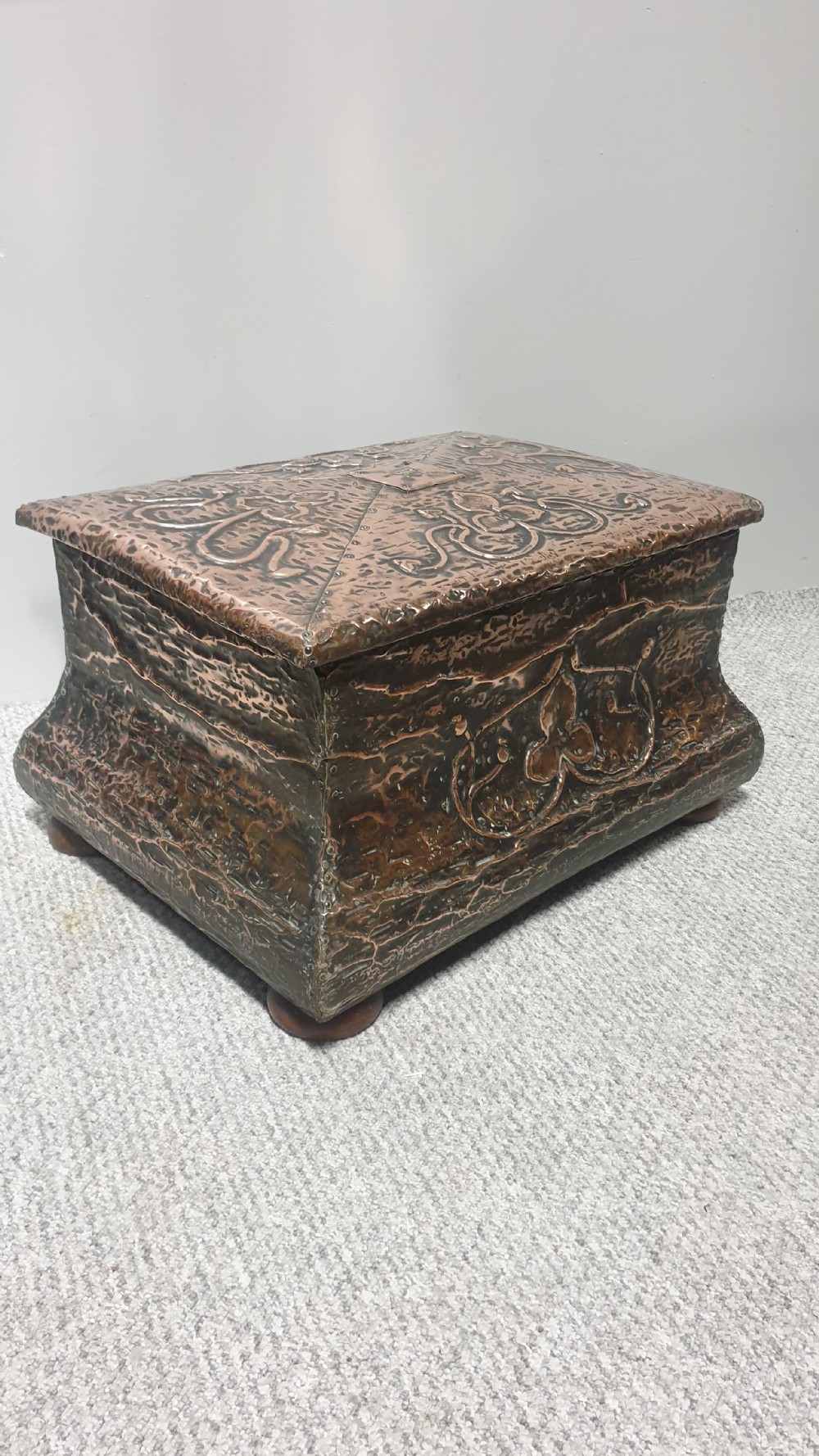 An antique rectangular copper fireside box featuring a hinged lid, hammered repousse foliate motifs, flared sides, and small bun feet. View V