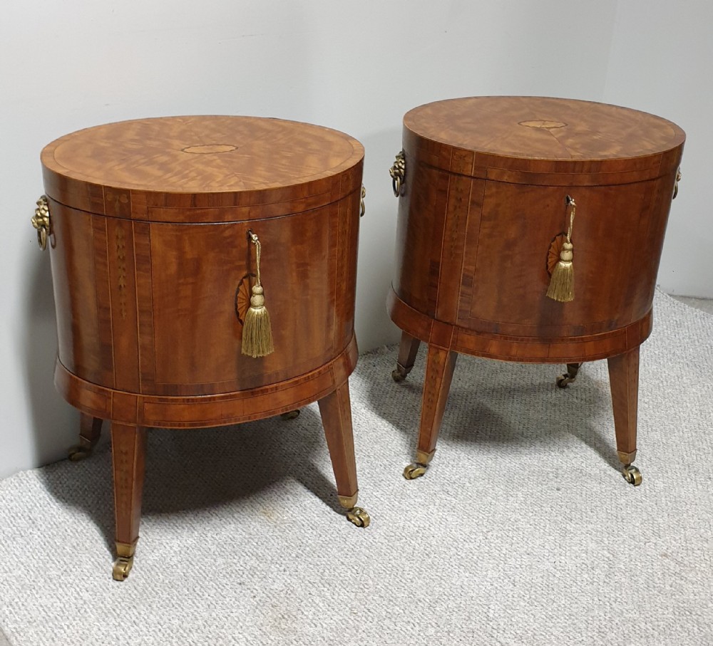 A pair of cylindrical satinwood wine coolers featuring inlaid marquetry oval medallions, lion mask brass side handles, key tassels, tapered legs, and brass castors.