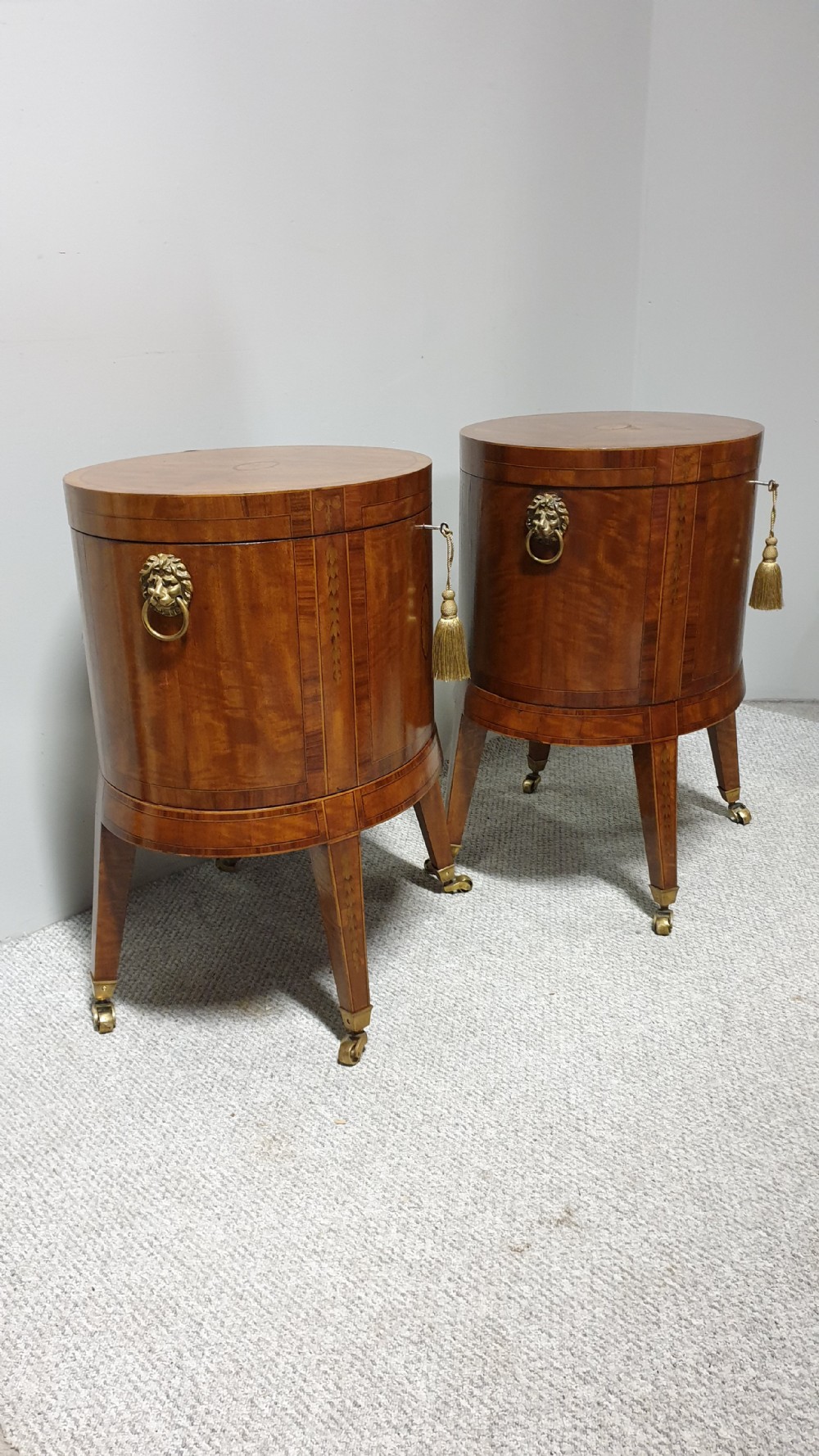 A pair of cylindrical satinwood wine coolers featuring inlaid marquetry oval medallions, lion mask brass side handles, key tassels, tapered legs, and brass castors. View IX