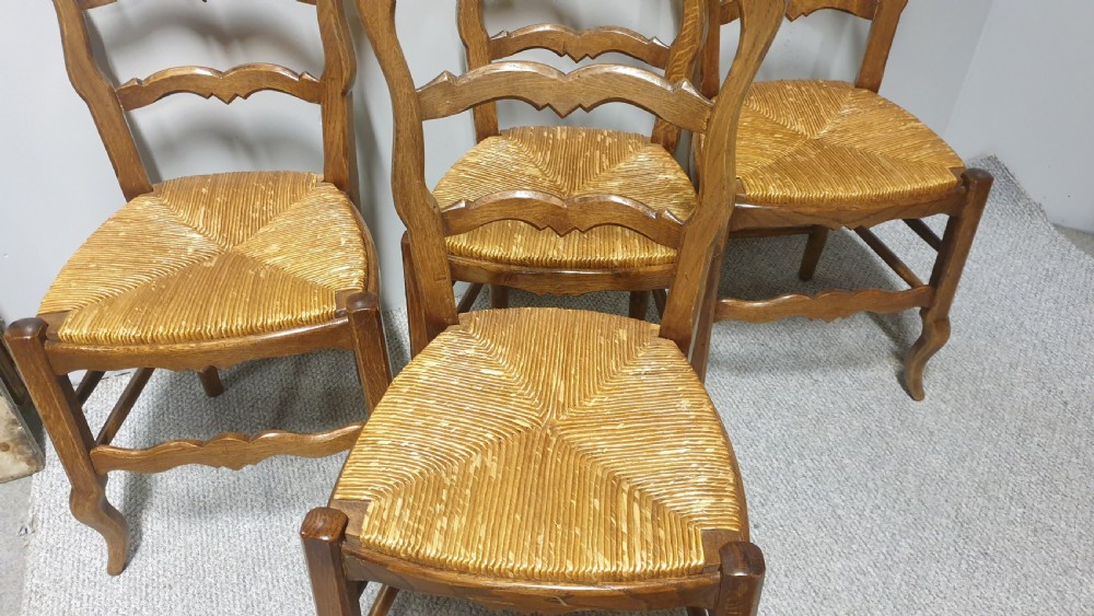 A set of four French Provincial oak dining chairs featuring carved floral top rails, wavy ladderbacks, woven rush seats, and cabriole front legs. View III