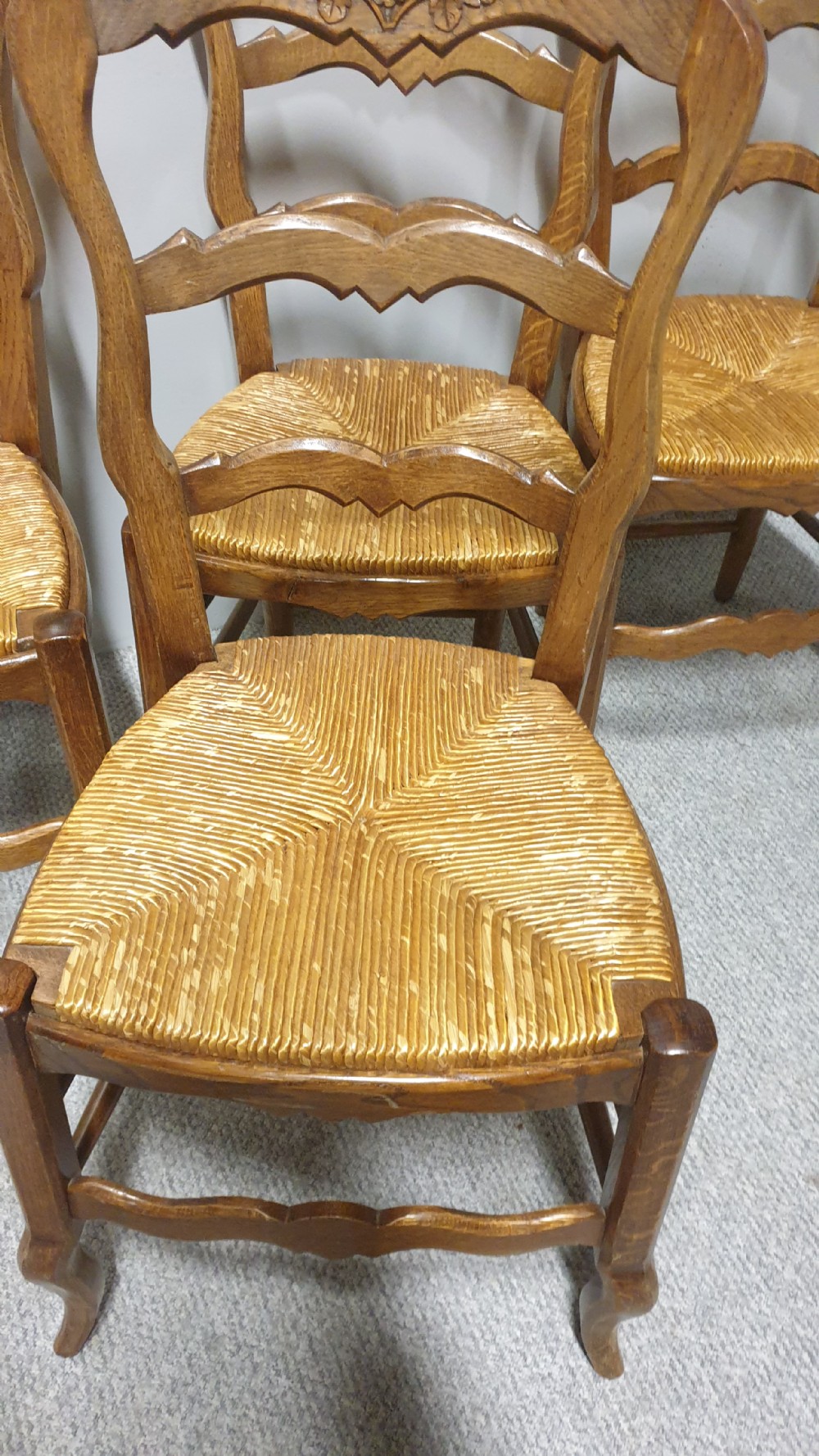 A set of four French Provincial oak dining chairs featuring carved floral top rails, wavy ladderbacks, woven rush seats, and cabriole front legs. View IV