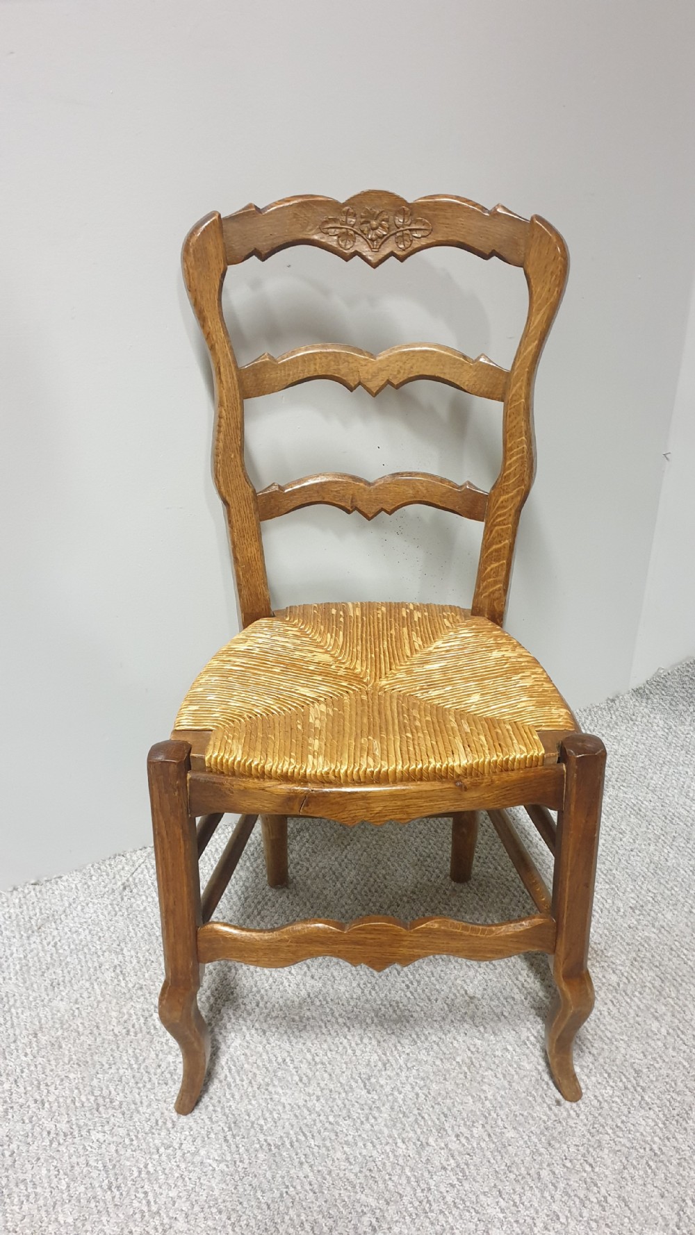 A set of four French Provincial oak dining chairs featuring carved floral top rails, wavy ladderbacks, woven rush seats, and cabriole front legs. View V
