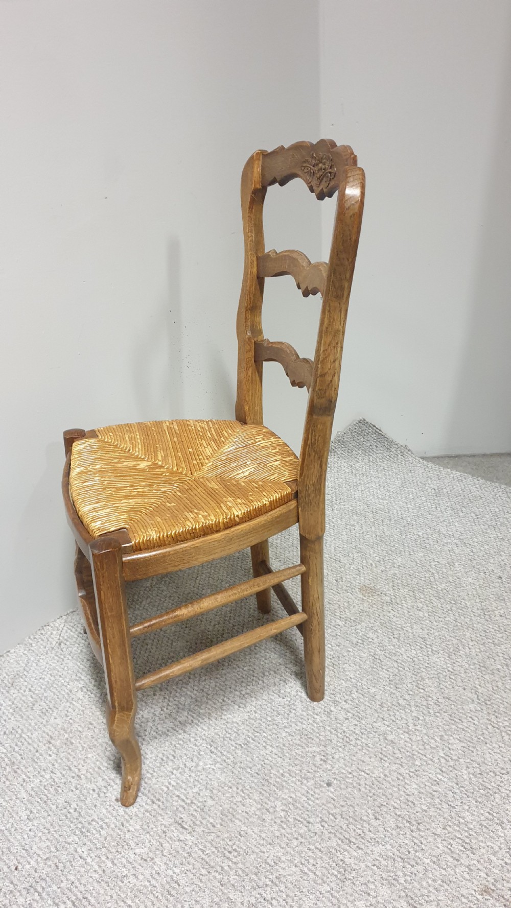 A set of four French Provincial oak dining chairs featuring carved floral top rails, wavy ladderbacks, woven rush seats, and cabriole front legs. View X