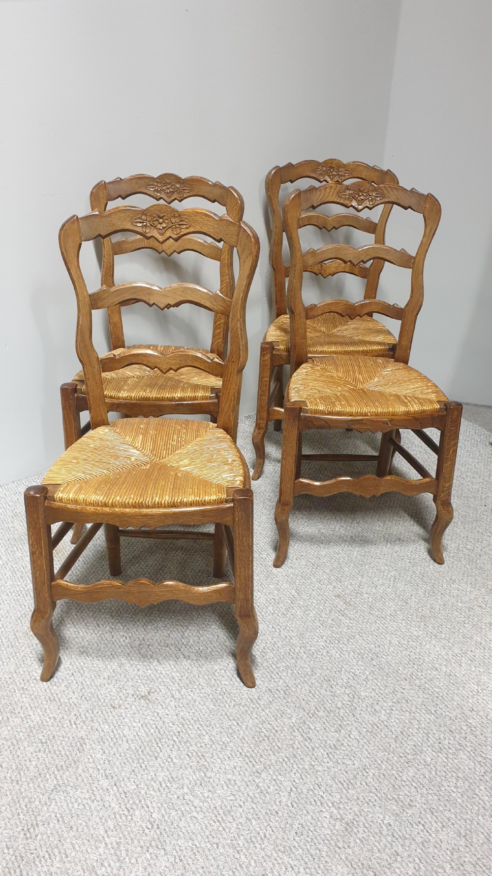 A set of four French Provincial oak dining chairs featuring carved floral top rails, wavy ladderbacks, woven rush seats, and cabriole front legs. View XII